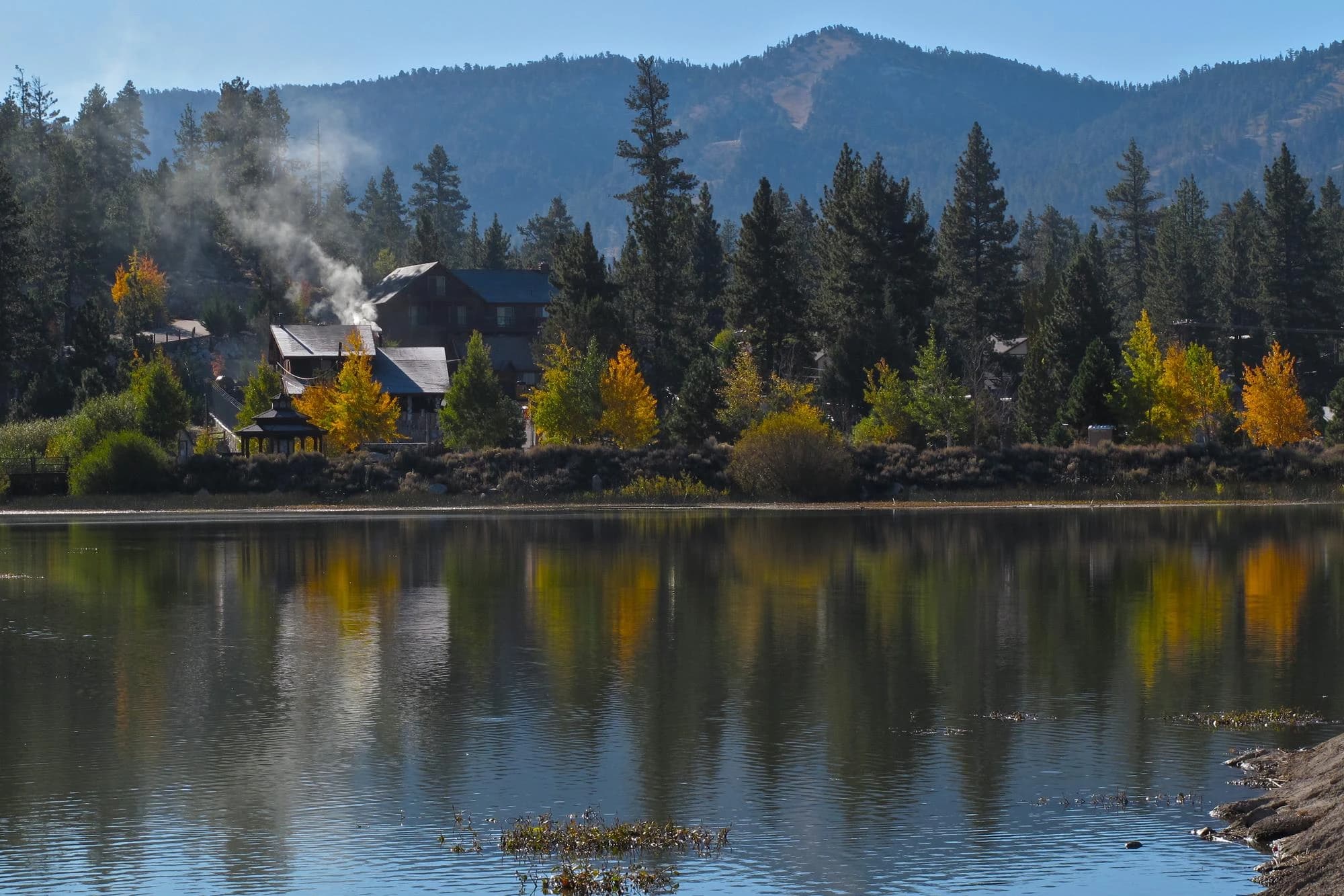 Autumn foliage reflecting in the still waters of Big Bear Lake, California, with a lakeside lodge and pine-covered mountains