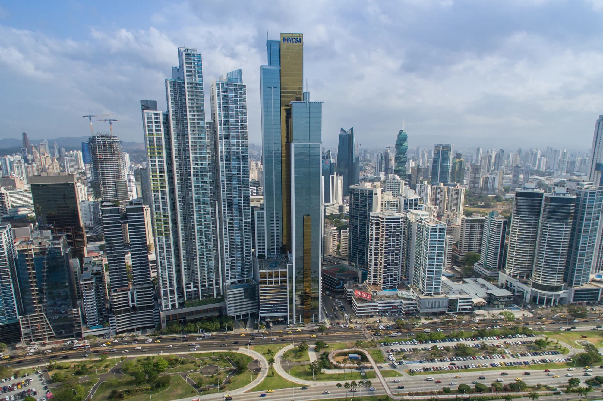 Aerial view of Panama City's dense downtown skyline of glass skyscrapers, including the gold-topped BICSA tower, with the Pacific coastline in the distance