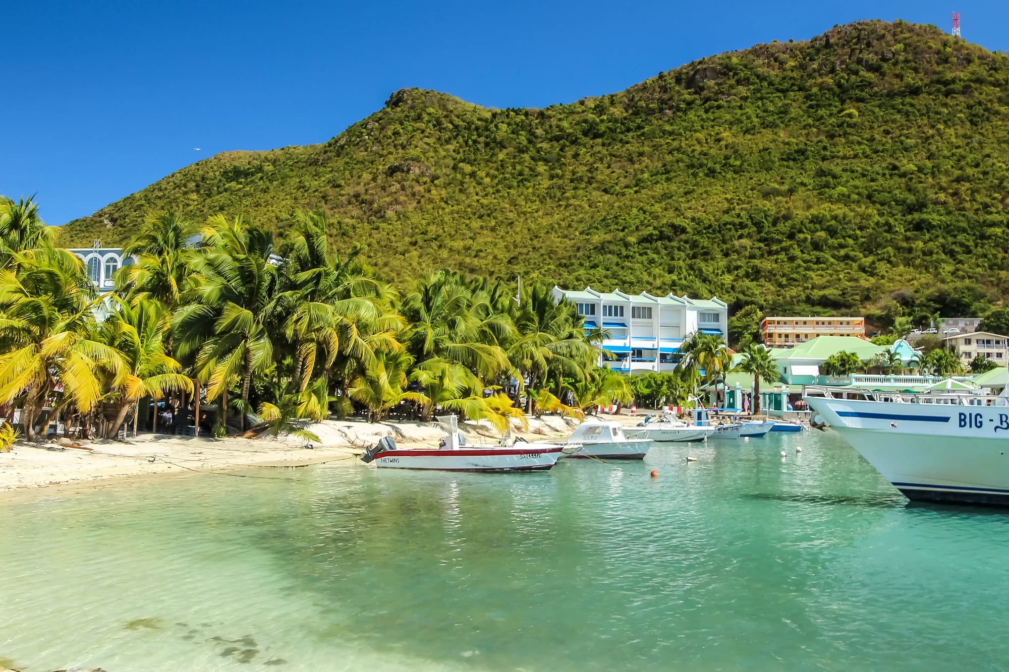 Great Bay Beach in Philipsburg, Sint Maarten, with palm trees, boats moored in turquoise water, and green hills rising behind the town